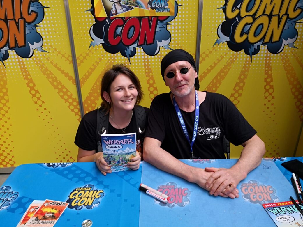 Zwei Personen sitzen an einem Tisch mit Comic-Con-Logo, die Frau hält ein Comic-Heft, der Mann trägt eine schwarze Mütze und Sonnenbrille.